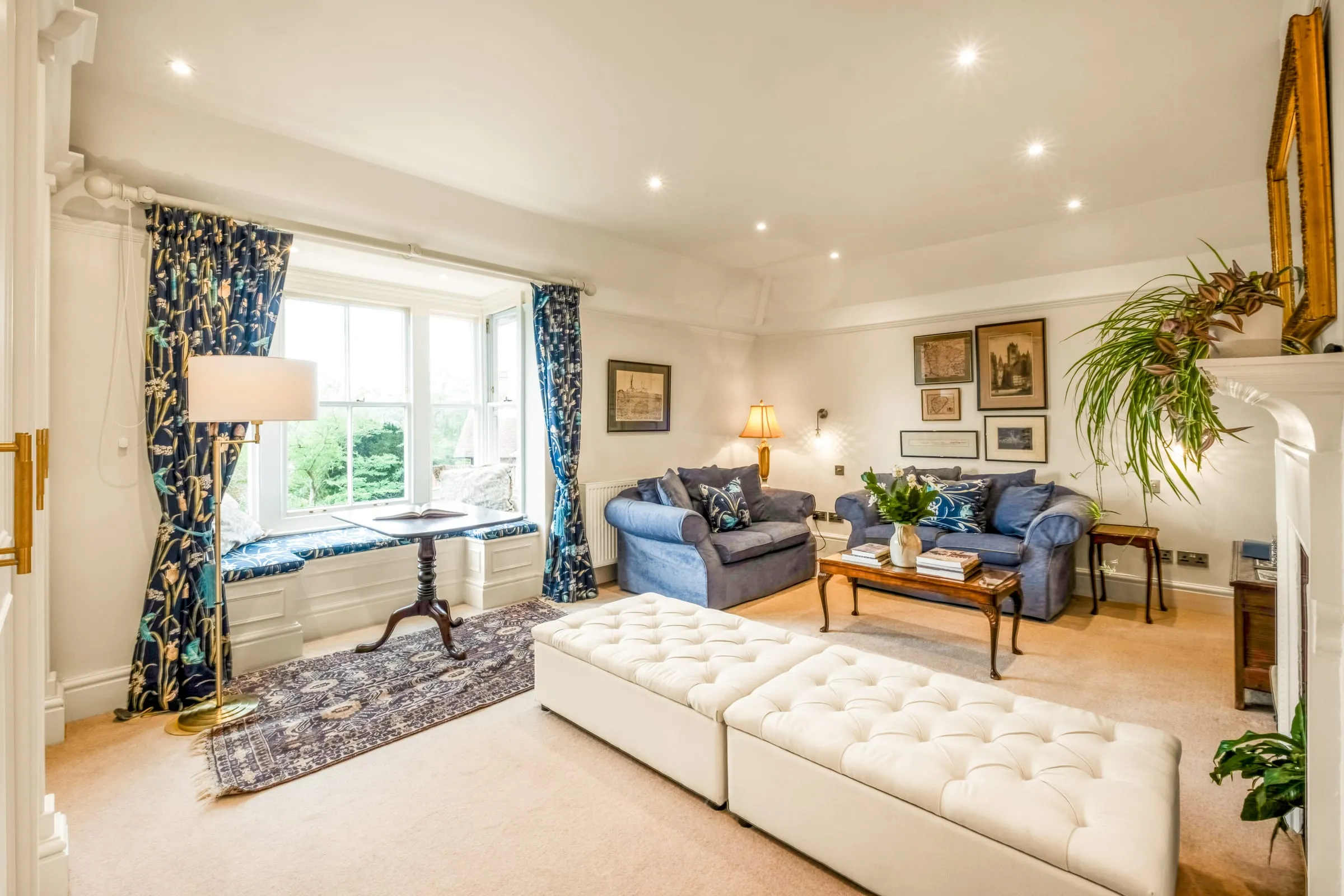 Living room with blue sofas, tufted cream ottoman and tall sash windows with patterned curtains